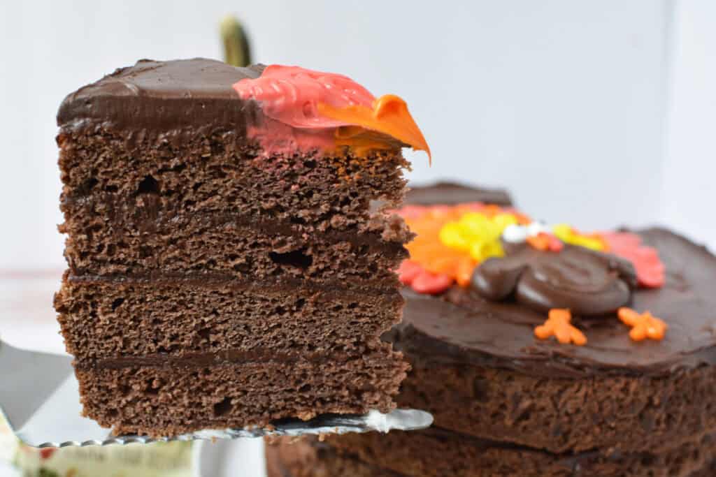 A close-up of a slice of Chocolate Thanksgiving Cake with rich chocolate frosting and colorful icing decorations, held above the remaining cake, which is also beautifully adorned with festive icing.
