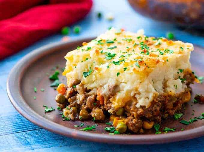 A serving of shepherds pie with a golden mashed potato topping sits on a brown plate, garnished with chopped parsley. The filling includes ground meat, peas, and diced carrots, making it one of those Easy Fall Dinner Ideas perfect for cozy evenings.