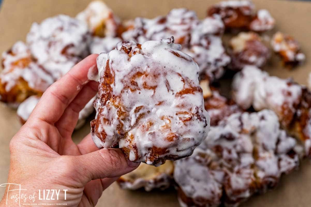 A hand holds a glazed apple fritter, with more fritters covered in white icing in the background on brown parchment paper.